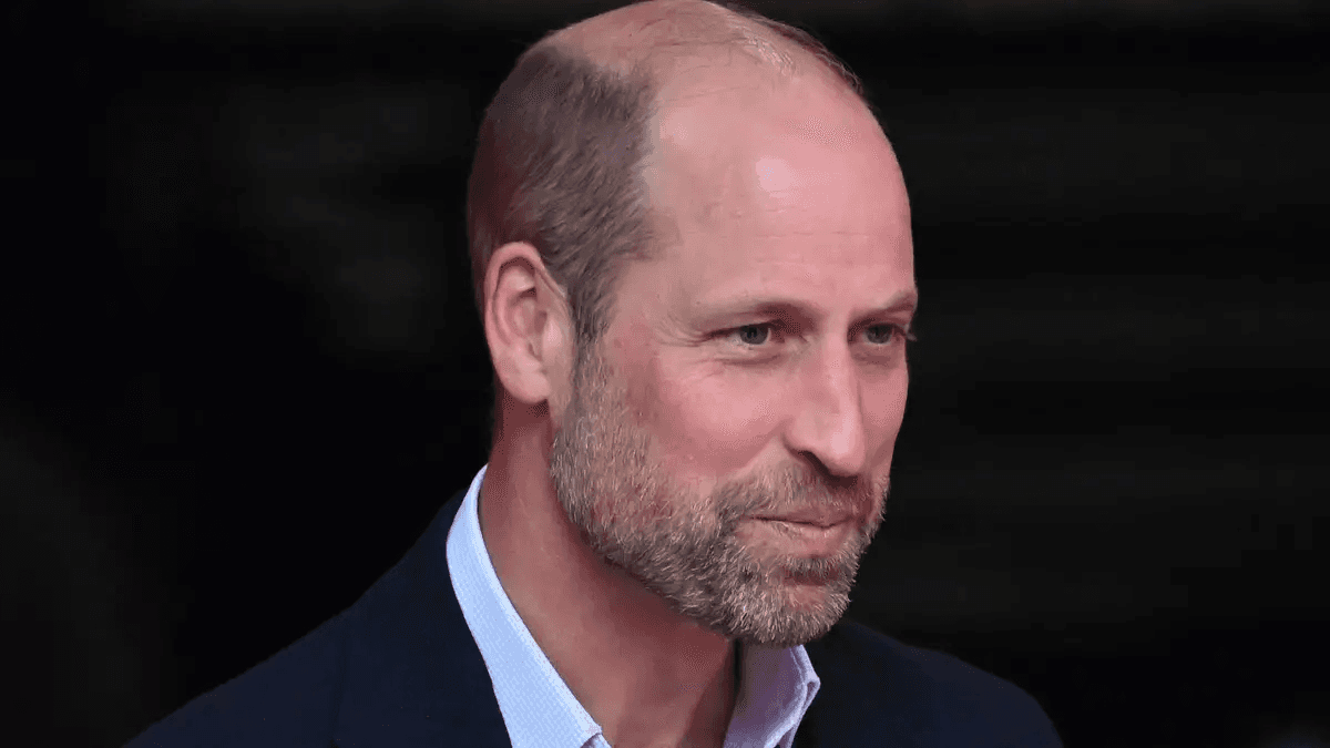 Prince William speaking at a public engagement, dressed in a navy suit, smiling warmly at the camera