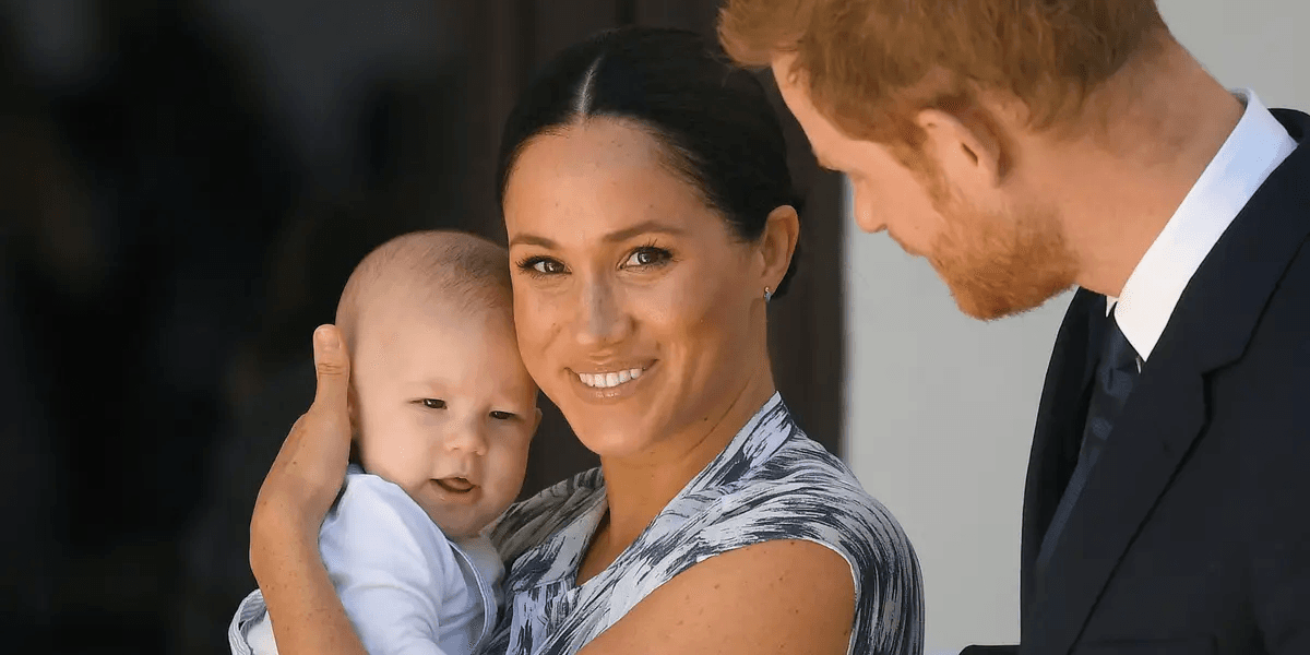 Meghan Markle and Prince Harry smiling together while Harry holds a young girl with long red hair, outdoors in natural light