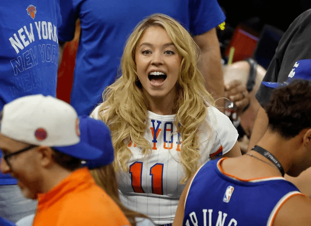 Sydney Sweeney wearing a white New York Knicks jersey and blue and orange jacket while sitting courtside at the NBA Finals