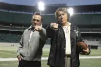 Two men in 1970s-era American football coaching attire stand on the sideline of a stadium, one large and broad-shouldered in a Raiders jacket, the other smaller and more wiry in a suit, both looking intently towards the field
