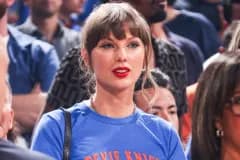 Taylor Swift smiling in the stands at an NBA game wearing a blue T-shirt reading Stevie Knicks, with the Haim sisters beside her in matching blue tops