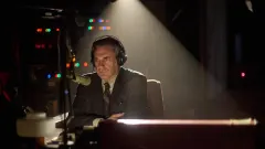 A man in 1970s clothing standing at a radio broadcasting desk against a dimly lit studio backdrop