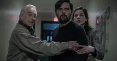 Three actors in a tense scene — a grey-haired older man on the left, a dark-haired man in the centre, and a woman on the right — appearing to confer urgently in what looks like an indoor setting