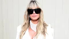 A woman with long wavy blonde hair wearing a lilac bikini and gold open-toe stilettos, carrying a large crochet tote bag and wearing dark sunglasses, photographed indoors in what appears to be a hotel room