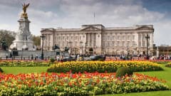 The ornate stone facade and gilded gates of a grand royal palace in London, with uniformed guards visible at the entrance