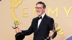 A bearded man in formal evening wear stands at an awards ceremony podium, holding an Emmy statuette and speaking into a microphone on a brightly lit stage