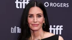 A woman with dark hair pulled back in a high ponytail wearing a black bikini and layered gold necklaces standing near ocean waves at a tropical beach resort