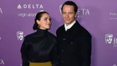 A woman with dark auburn hair wearing an elegant outfit smiles at a formal awards event, standing beside a man in a black tuxedo