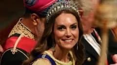 A woman in a flowing buttercup yellow gown with a sheer cape, wearing a diamond and pearl tiara, standing at a formal royal dinner setting inside a grand castle room