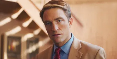 A man in a dark suit stands in what appears to be a television studio setting, looking directly ahead with an intense expression