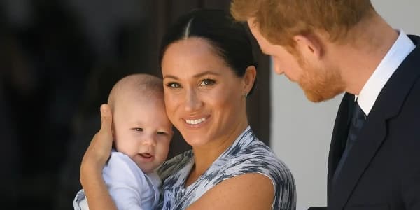 Meghan Markle and Prince Harry smiling together while Harry holds a young girl with long red hair, outdoors in natural light