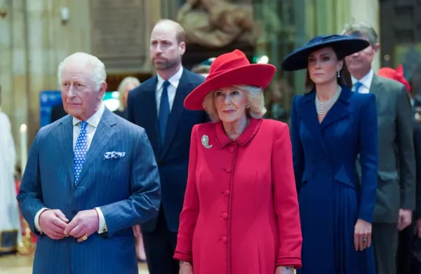 A formal royal gathering showing a man in a dark suit standing at a podium, with ornate palace interiors visible in the background