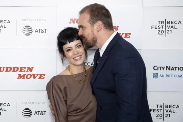 David Harbour speaking at a press event, wearing a dark suit and smiling at the camera