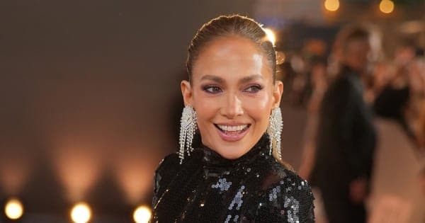 Jennifer Lopez smiling and raising her arms in celebration, wearing a mint green skirt and beige top while watching television