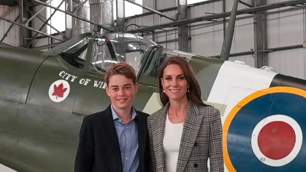 A young boy wearing a navy blue suit jacket and blue collared shirt standing on an airfield against a backdrop of military aircraft hangars, accompanied by a woman in a tweed jacket.