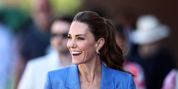 A woman wearing a blue linen trouser suit with her hair in a ponytail, greeting members of the public against a backdrop of the All England Club at Wimbledon.