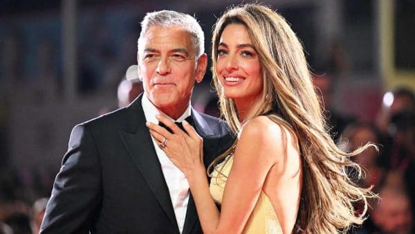 A man in a grey suit and a woman in a black embroidered dress stepping off a boat onto a lakeside dock, against a scenic Italian lake backdrop.