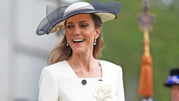 A woman in an elegant coat dress waves to a crowd at a formal public engagement, her dark hair neatly styled