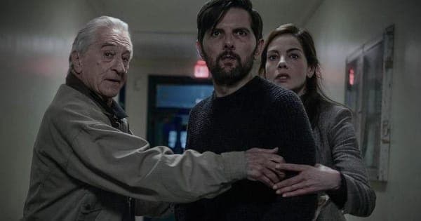 Three actors in a tense scene — a grey-haired older man on the left, a dark-haired man in the centre, and a woman on the right — appearing to confer urgently in what looks like an indoor setting