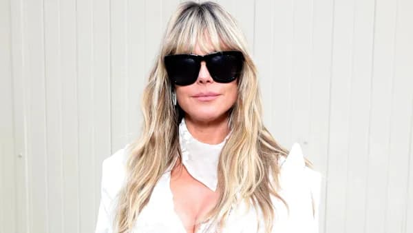 A woman with long wavy blonde hair wearing a lilac bikini and gold open-toe stilettos, carrying a large crochet tote bag and wearing dark sunglasses, photographed indoors in what appears to be a hotel room