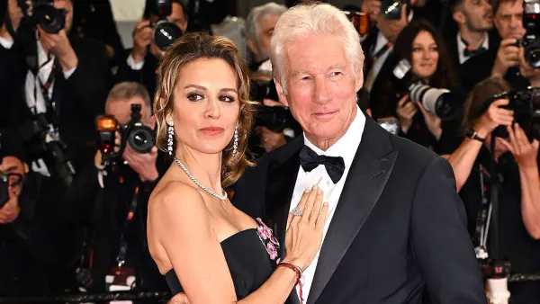 A silver-haired man in casual evening wear stands with a woman in a white maxi dress and block heels at an intimate outdoor birthday gathering, both smiling warmly