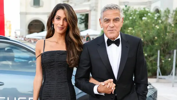 A woman in a sleek black pleated gown with leather details stands on a red carpet at a film festival, her brunette hair styled in soft tousled waves. A man in a dark suit stands beside her.