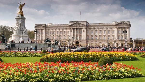 The ornate stone facade and gilded gates of a grand royal palace in London, with uniformed guards visible at the entrance