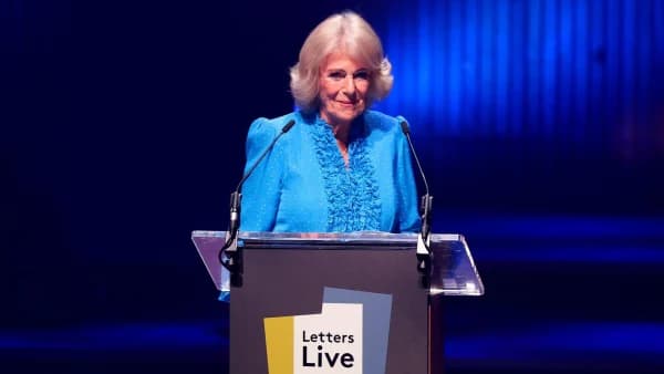 A woman in a blue evening gown stands at a lectern on a stage at a large London concert hall, addressing a packed audience