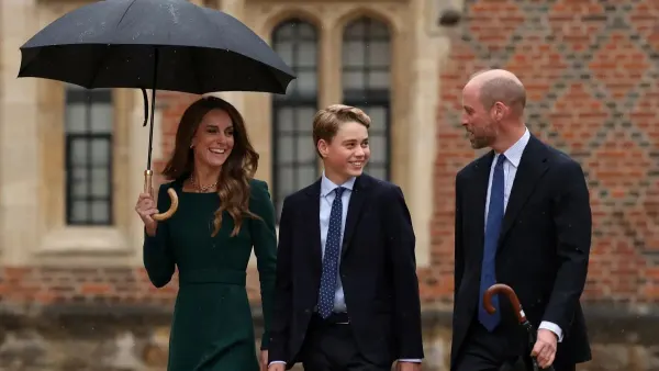 A teenage boy in a dark blazer, shirt, tie and chinos walks across a cobblestone courtyard with two adults beside him in the rain, approaching a historic stone building