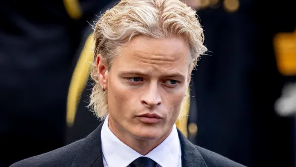 A young man in a dark suit walking into a grand cathedral alongside other formally dressed individuals, with stone pillars and floral arrangements visible in the background