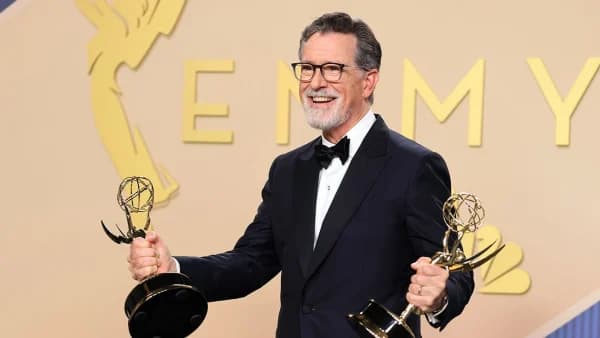 A bearded man in formal evening wear stands at an awards ceremony podium, holding an Emmy statuette and speaking into a microphone on a brightly lit stage