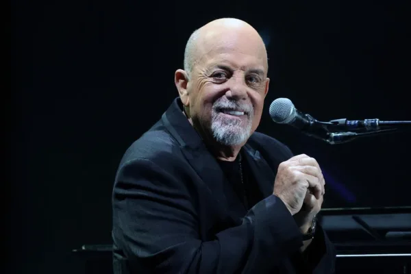 An older man with grey hair in a dark jacket speaking into a microphone on stage, hands raised, performing in front of a large audience