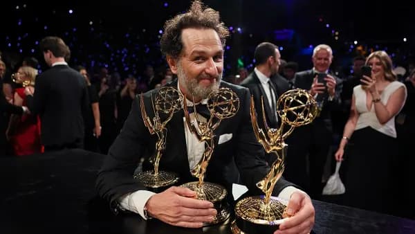 A man in formal evening wear holding two Emmy statuettes on a stage at an awards ceremony, smiling at the crowd