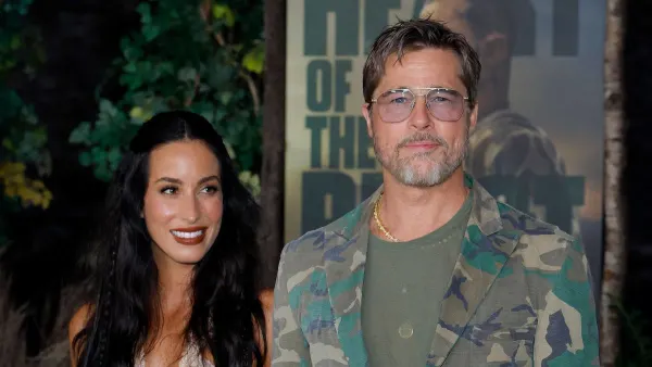 A man in a camouflage suit and aviator sunglasses and a woman in a pale pink tiered dress with lace trim walk hand-in-hand on a red carpet at an outdoor premiere venue