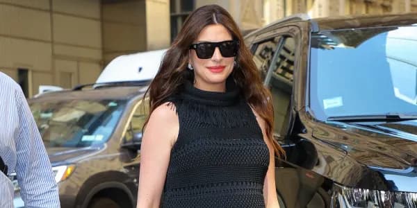 A woman with dark hair wearing a black crochet maxi dress with fringe detailing at the neckline and hem, carrying a black leather handbag decorated with colourful beaded embellishments, photographed outdoors in an urban setting