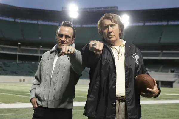 Two men in 1970s-era American football coaching attire stand on the sideline of a stadium, one large and broad-shouldered in a Raiders jacket, the other smaller and more wiry in a suit, both looking intently towards the field
