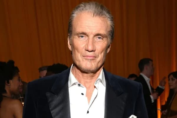 A broad-shouldered man with cropped grey hair, wearing a dark jacket, photographed at a media event against a branded backdrop