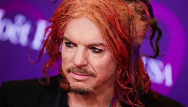 A man with curly red hair standing on a stage in front of a colourful backdrop, wearing a dark outfit
