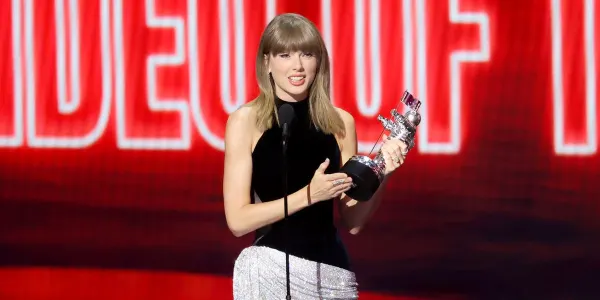 A woman in an elaborate stage outfit stands at a podium on a large awards show stage, holding a trophy and speaking into a microphone, surrounded by dramatic lighting