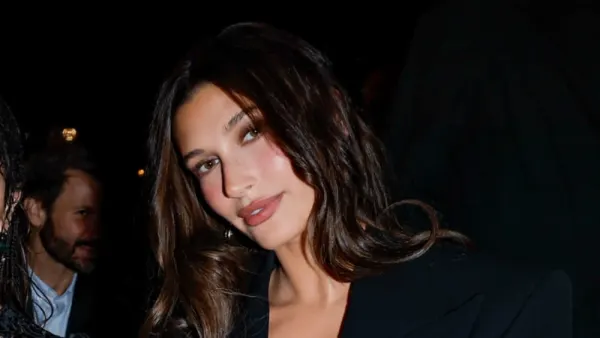 A woman with a short dark brunette bob sits front row at a fashion show wearing a plunging black suit coat, layered rings and narrow dark sunglasses