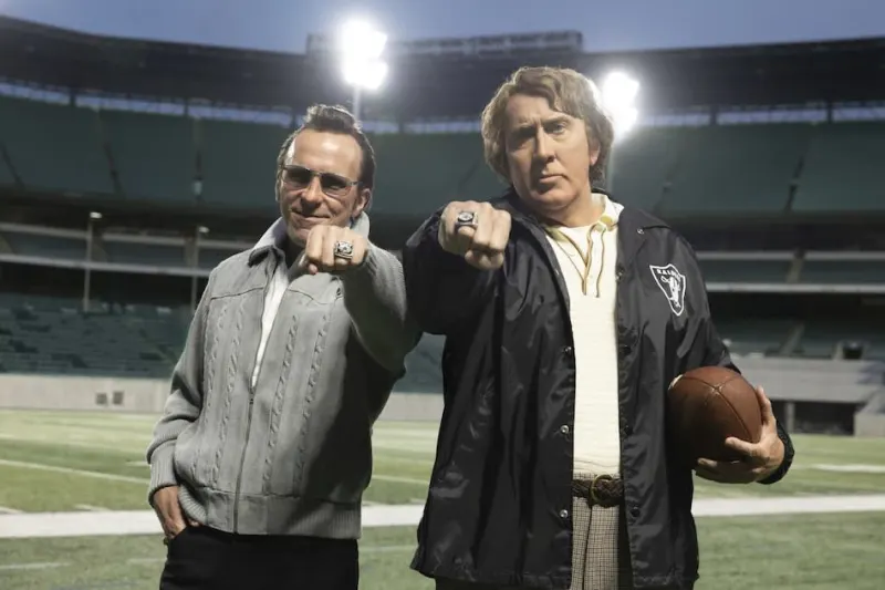Two men in 1970s-era American football coaching attire stand on the sideline of a stadium, one large and broad-shouldered in a Raiders jacket, the other smaller and more wiry in a suit, both looking intently towards the field