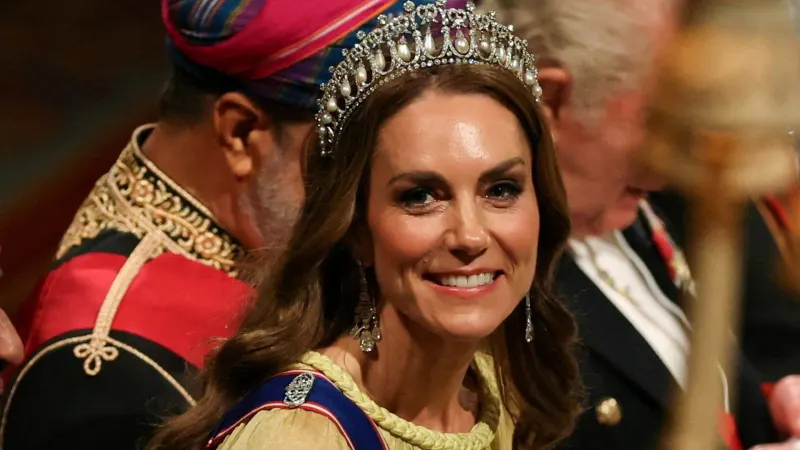 A woman in a flowing buttercup yellow gown with a sheer cape, wearing a diamond and pearl tiara, standing at a formal royal dinner setting inside a grand castle room