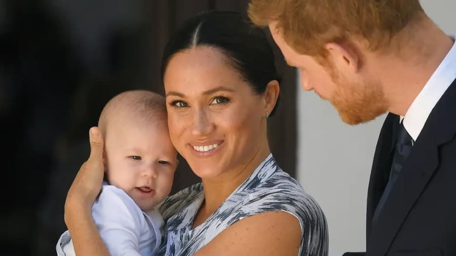 Meghan Markle and Prince Harry smiling together while Harry holds a young girl with long red hair, outdoors in natural light