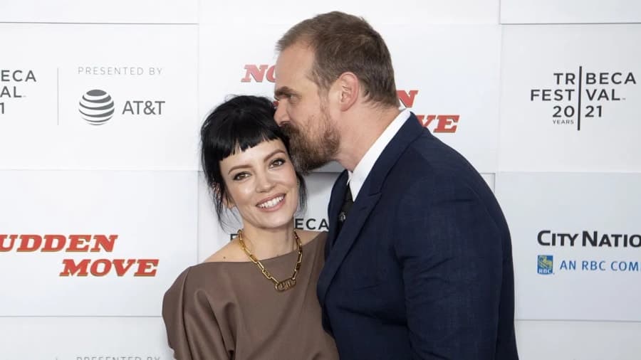 David Harbour speaking at a press event, wearing a dark suit and smiling at the camera