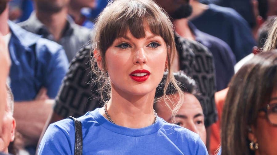Taylor Swift smiling in the stands at an NBA game wearing a blue T-shirt reading Stevie Knicks, with the Haim sisters beside her in matching blue tops