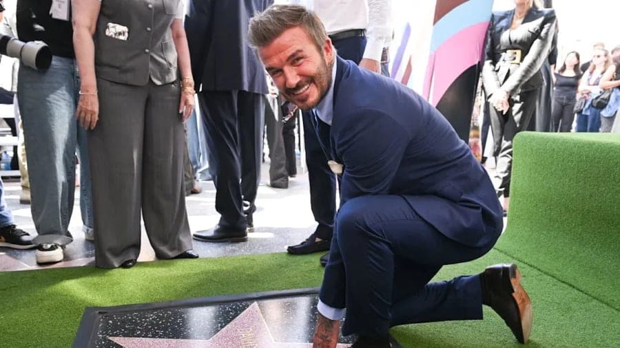 David Beckham smiling at his Hollywood Walk of Fame star ceremony, surrounded by family and friends including Tom Cruise, with a green football-themed carpet visible