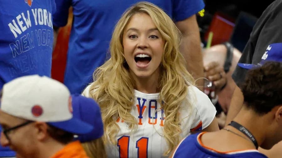 Sydney Sweeney wearing a white New York Knicks jersey and blue and orange jacket while sitting courtside at the NBA Finals