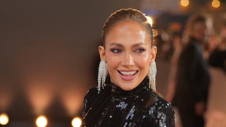 Jennifer Lopez smiling and raising her arms in celebration, wearing a mint green skirt and beige top while watching television