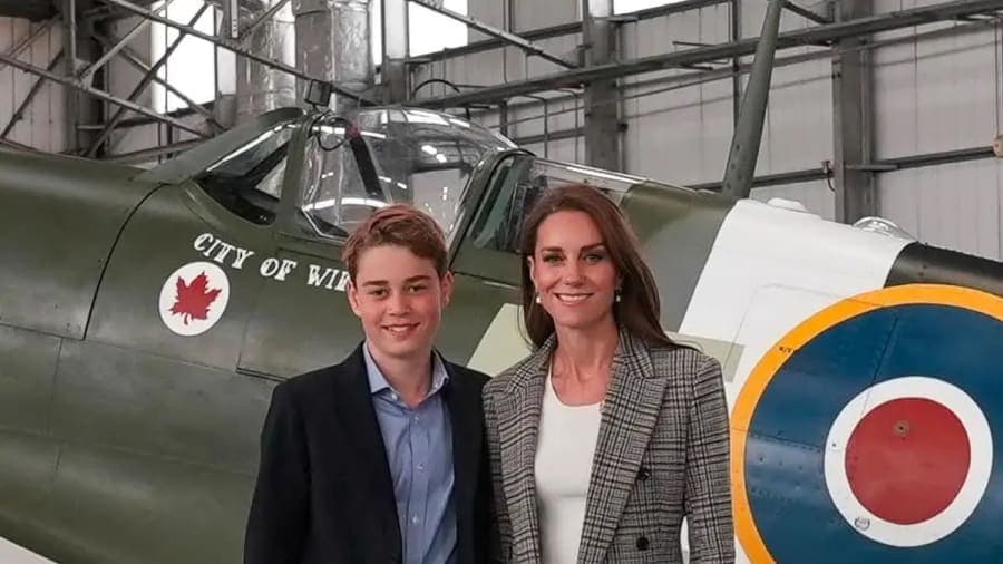 A young boy wearing a navy blue suit jacket and blue collared shirt standing on an airfield against a backdrop of military aircraft hangars, accompanied by a woman in a tweed jacket.