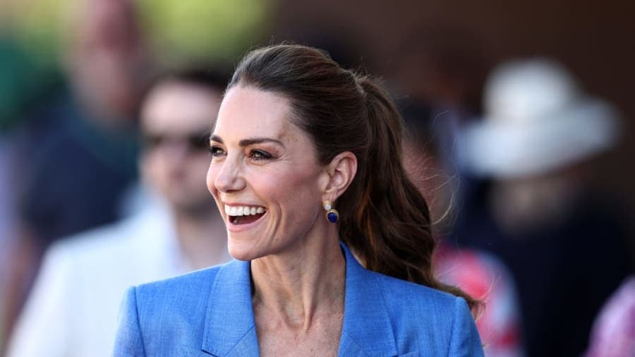 A woman wearing a blue linen trouser suit with her hair in a ponytail, greeting members of the public against a backdrop of the All England Club at Wimbledon.